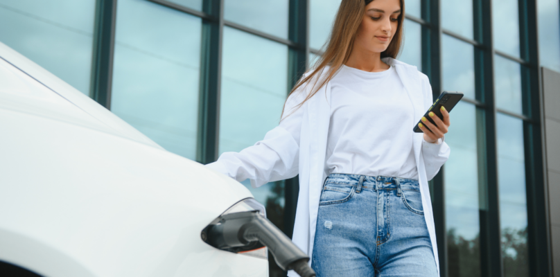 A woman waiting while her electric vehicle charges is representative of auto financing.