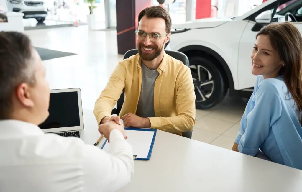 A man shakes hands with a salesman at a dealership while sitting next to a woman.