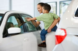 A woman carries her toddler aged son while looking at a sticker price of a car at a dealership