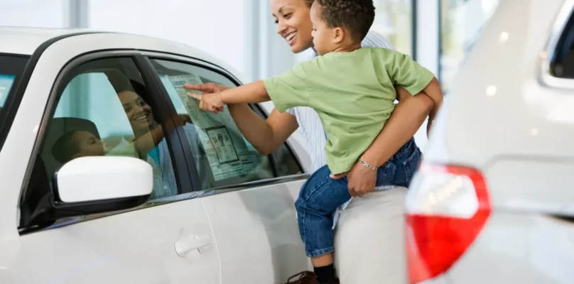 A woman carries her toddler aged son while looking at a sticker price of a car at a dealership